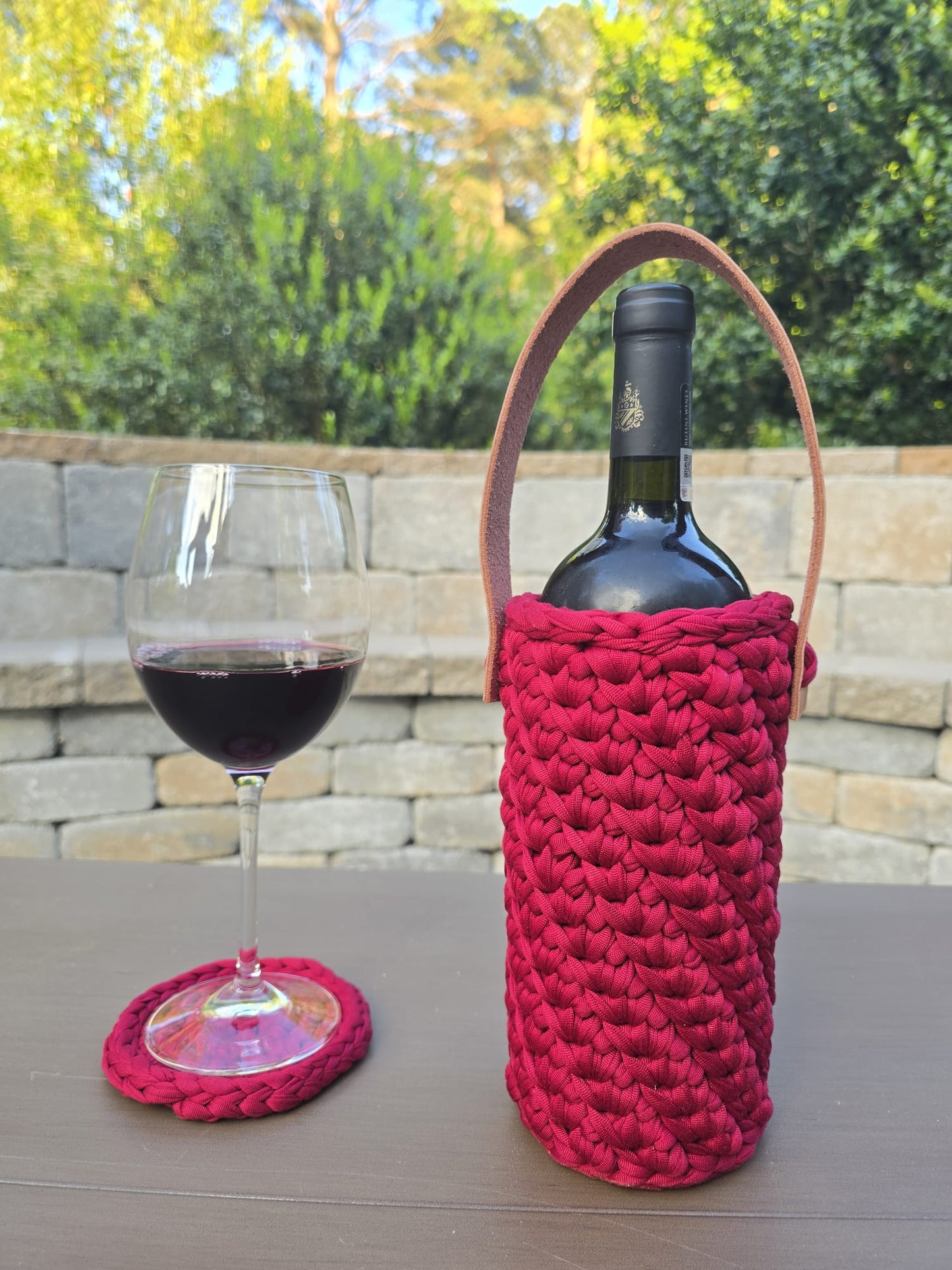 Handmade crochet wine holder with leather handle displayed outdoors beside a glass of red wine – eco-friendly gift for wine lovers.