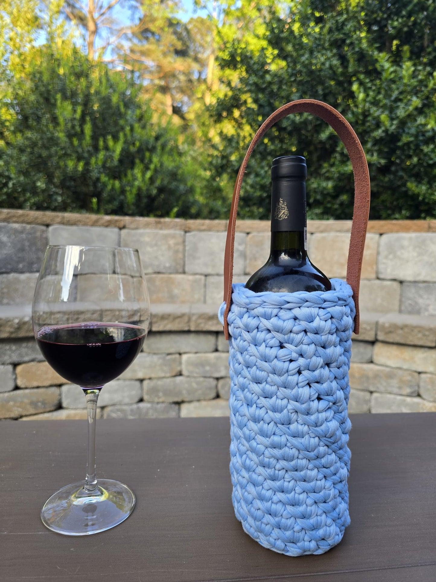 Handmade crochet wine holder with leather handle displayed outdoors beside a glass of red wine – eco-friendly gift for wine lovers.