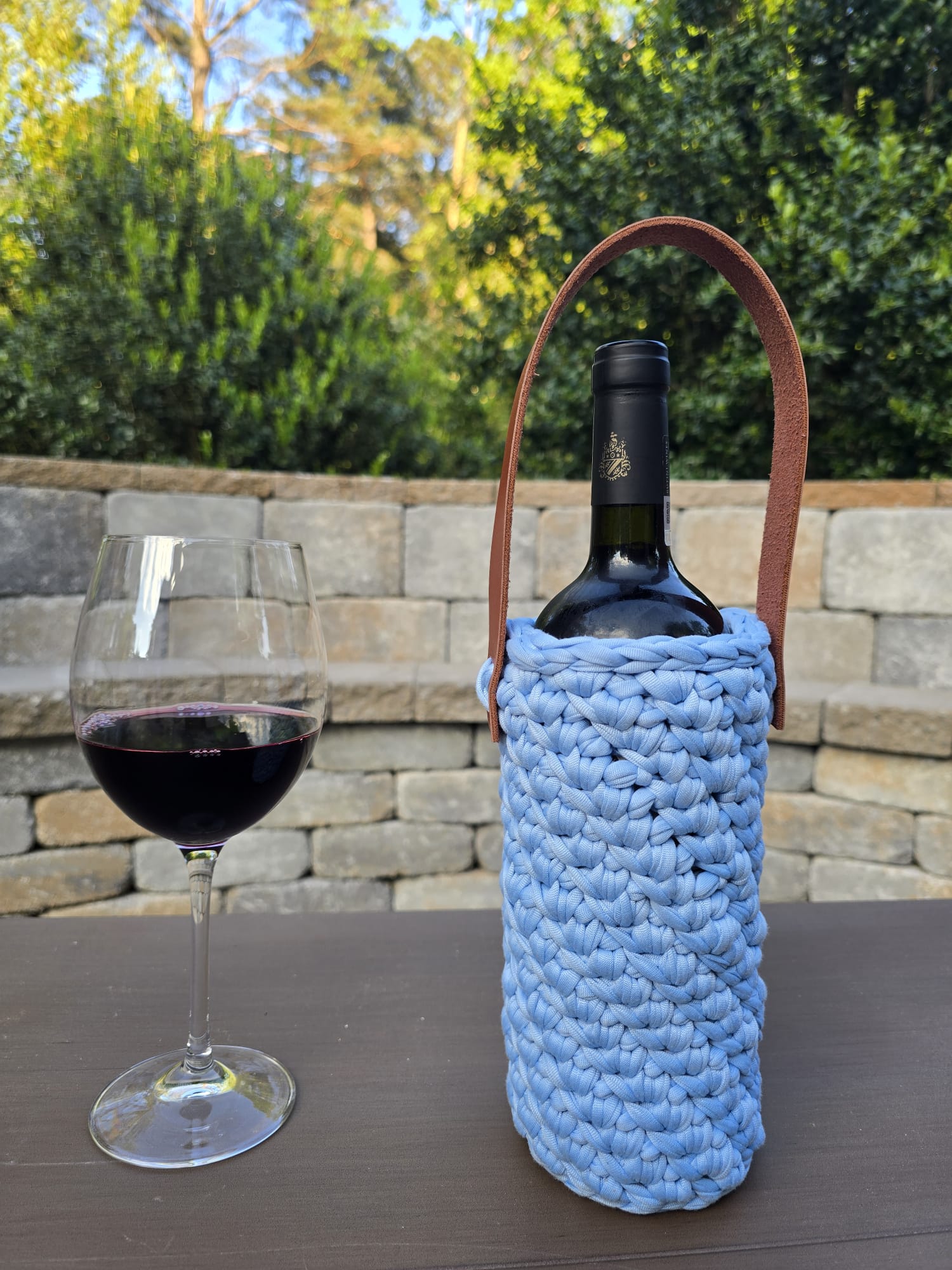 Handmade crochet wine holder with leather handle displayed outdoors beside a glass of red wine – eco-friendly gift for wine lovers.