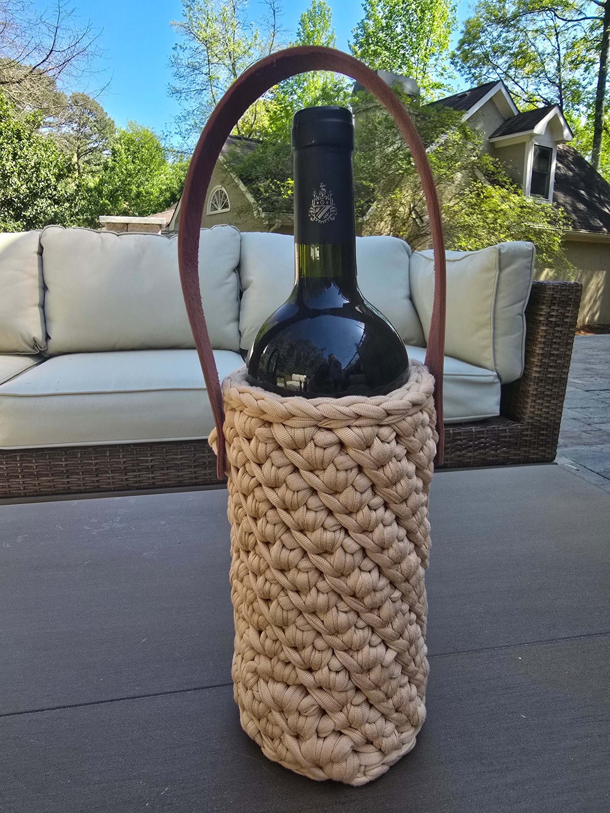 Handmade crochet wine holder with leather handle displayed outdoors beside a glass of red wine – eco-friendly gift for wine lovers.