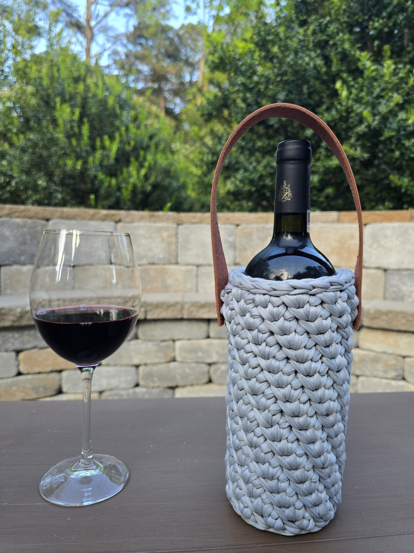 Handmade crochet wine holder with leather handle displayed outdoors beside a glass of red wine – sustainable gift for wine lovers.